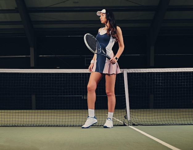 Giocatore di tennis femminile castana sexy che posa su un campo da tennis dell'interno.