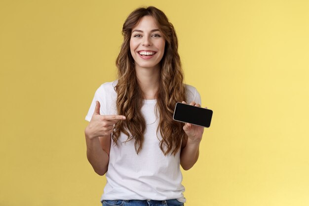 Giocatore femminile spensierato carismatico carino dai capelli ricci ama giocare a giochi per smartphone che mostrano il proprio punteggio di gioco tenere il telefono cellulare orizzontale che punta display ridendo contento divertito. Copia spazio