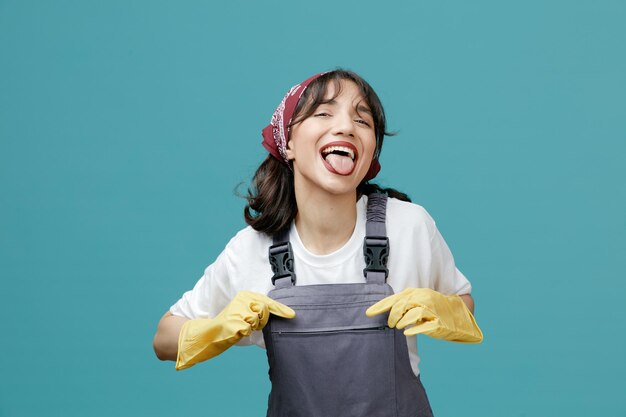 Giocosa giovane donna addetta alle pulizie che indossa bandana uniforme e guanti di gomma che afferrano la sua uniforme guardando la fotocamera che mostra la lingua isolata su sfondo blu