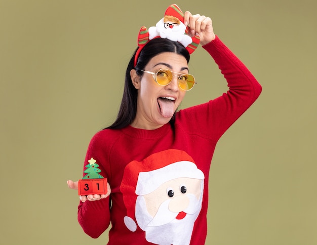 Giocosa giovane donna caucasica che indossa la fascia santa e maglione con gli occhiali che tengono il giocattolo dell'albero di Natale