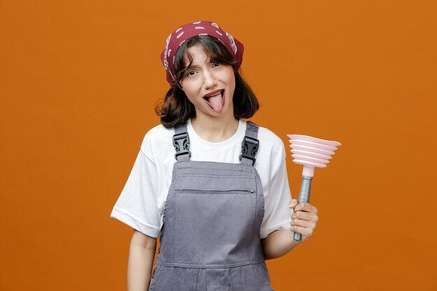 Giocosa giovane donna detergente che indossa uniforme e bandana tenendo lo stantuffo guardando la fotocamera che mostra la lingua isolata su sfondo arancione