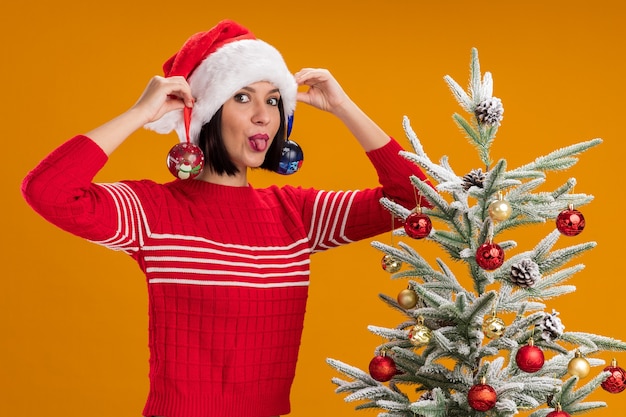 Giocosa ragazza che indossa il cappello di Babbo Natale in piedi vicino all'albero di Natale decorato appeso palline di Natale sulle orecchie guardando la telecamera che mostra la lingua isolata su sfondo arancione