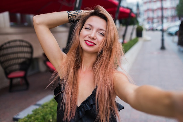 Giocoso carino rosso sente donna con sorridente che fa autoritratto e godersi le vacanze estive in Europa. Immagine esterna positiva. Abito nero, labbra rosse.
