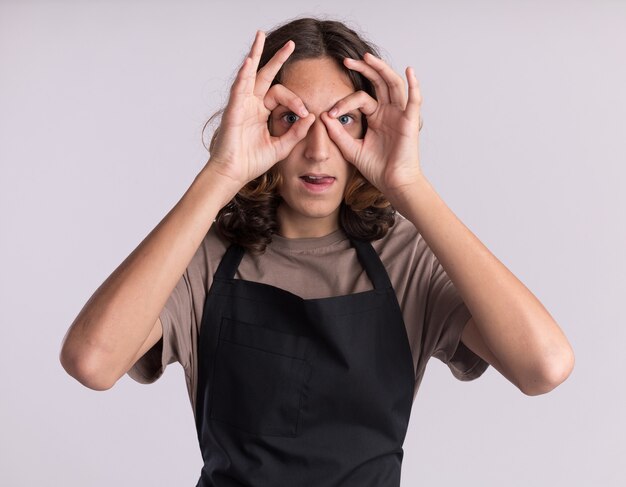 Giocoso giovane barbiere bello che indossa l'uniforme che fa un gesto di sguardo usando le mani come binocolo che mostra la lingua