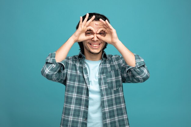 giocoso giovane bell'uomo che guarda la fotocamera che mostra il gesto del binocolo sorridente isolato su sfondo blu