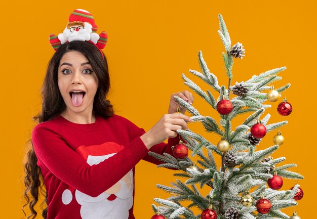 Giocoso giovane bella ragazza che indossa la fascia di Babbo Natale e maglione in piedi vicino all'albero di Natale decorarlo guardando la telecamera che mostra la linguetta isolata su sfondo arancione