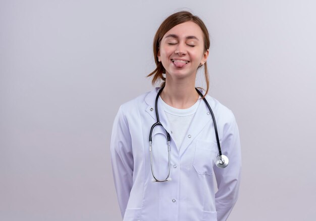 Giocoso giovane dottoressa indossa veste medica e stetoscopio che mostra la lingua con gli occhi chiusi con lo spazio della copia