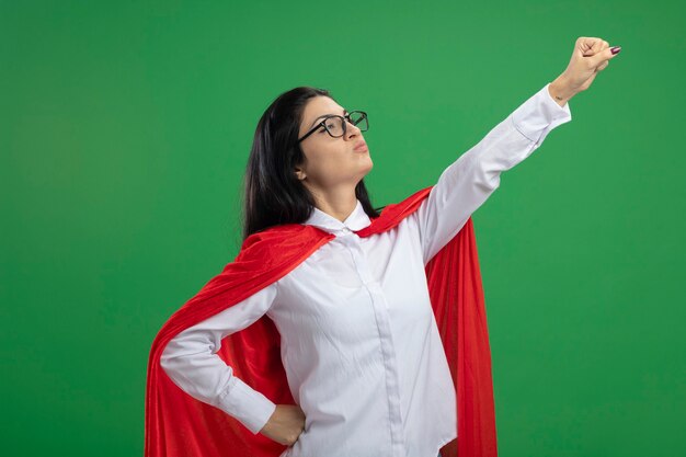 Giocoso giovane supereroe caucasico ragazza con gli occhiali in piedi in superman posa in vista di profilo alzando il pugno isolato su sfondo verde con spazio di copia