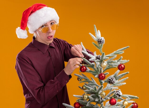 Giocoso giovane uomo biondo che indossa cappello santa e occhiali in piedi in vista di profilo vicino albero di Natale decorato su sfondo arancione