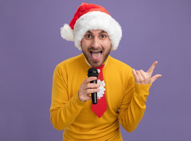 Giocoso giovane uomo caucasico indossando il cappello di Natale e cravatta tenendo il microfono guardando la telecamera che mostra la lingua facendo segno di roccia isolato su sfondo viola con spazio di copia