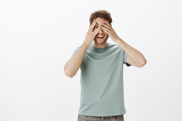 giocoso modello di uomo felice in t-shirt, che copre gli occhi con i palmi delle mani e sbirciare tra le dita, sorridendo ampiamente