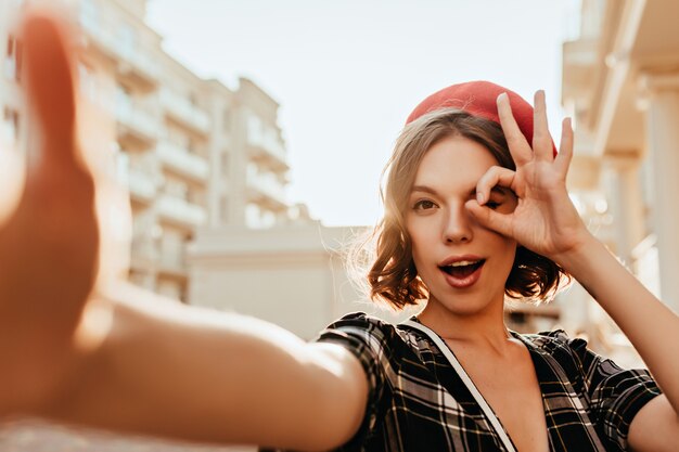 Giocoso signora francese in posa sulla strada. Ragazza caucasica in berretto elegante scherzare mentre fa selfie.