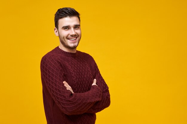 Gioia, felicità e reazione positiva. Uomo europeo carismatico bello con un ampio sorriso radioso che posa al muro giallo vuoto con lo spazio della copia per il contenuto pubblicitario, tenendo le braccia conserte