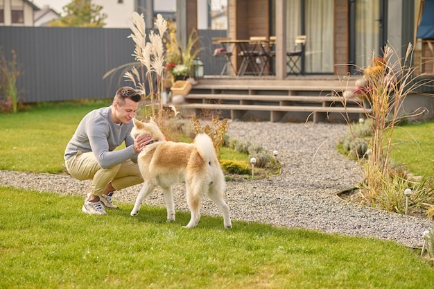 Gioia reciproca. Giovane adulto sorridente in abiti casual accovacciato toccando guardando il suo cane a piedi vicino alla casa di campagna nel bel pomeriggio