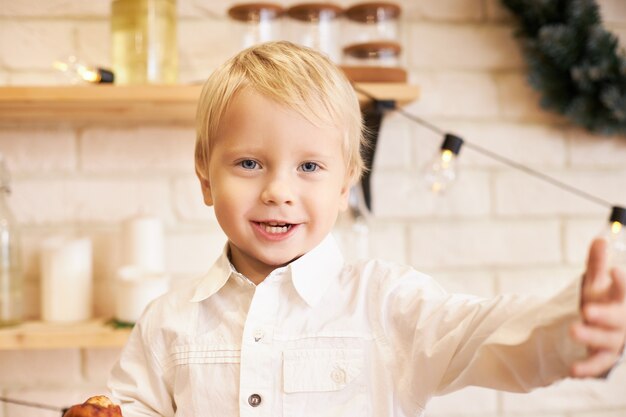 Gioia, svago ed emozioni positive. Ritratto di ragazzino sveglio emotivo in camicia bianca gesticolando attivamente essendo affamato, andando a fare uno spuntino prima di pranzo, dicendo qualcosa, in posa in cucina accogliente