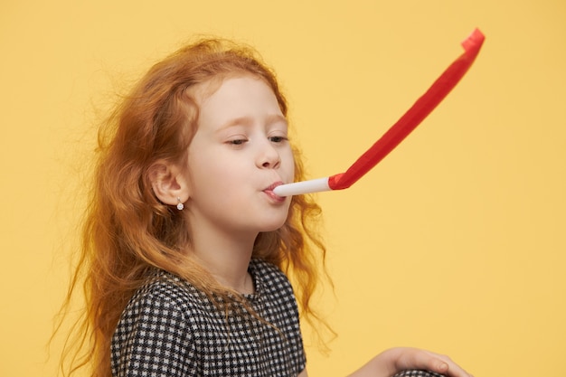 Gioiosa bambina allegra con lunghi capelli rossi che soffia il corno del partito