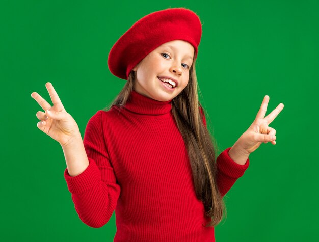 Gioiosa bambina bionda che indossa un berretto rosso guardando la parte anteriore che mostra il segno di pace isolato sul muro verde