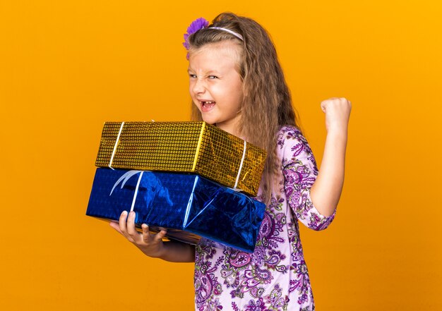 gioiosa bambina bionda con scatole regalo e tenendo il pugno isolato sulla parete arancione con spazio di copia