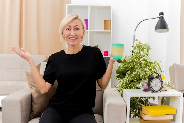 Gioiosa bella donna russa bionda si siede sulla poltrona tenendo la tazza e guardando