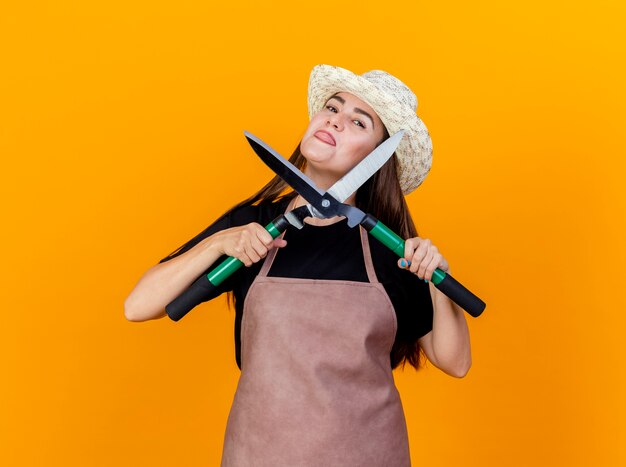 Gioiosa bella ragazza giardiniere che indossa uniforme e cappello da giardinaggio tenendo i clippers e mostrando la lingua