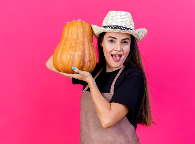 Gioiosa bella ragazza giardiniere in uniforme che indossa cappello da giardinaggio tenendo la zucca intorno al viso isolato su sfondo rosa