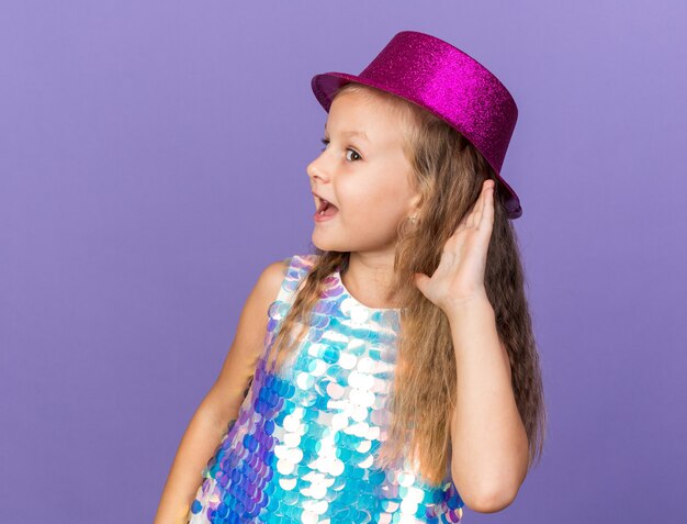 gioiosa bimba bionda con cappello da festa viola che tiene la mano vicino all'orecchio cercando di sentire isolato sul muro viola con spazio copia