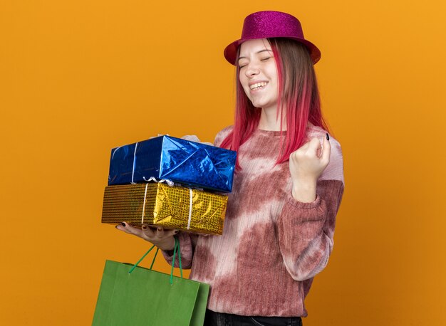 Gioiosa con gli occhi chiusi giovane bella ragazza che indossa un cappello da festa che tiene in mano una borsa regalo con scatole regalo che mostrano un gesto di sì