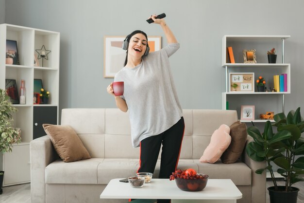 Gioiosa con gli occhi chiusi ragazza che indossa le cuffie che tiene il telecomando della tv con una tazza di tè in piedi dietro il tavolino da caffè in soggiorno