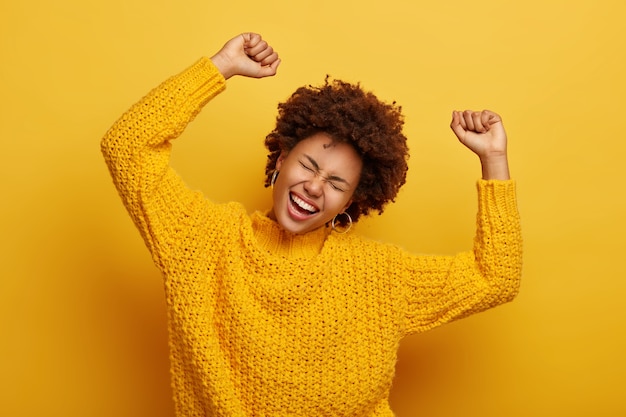 Gioiosa donna afro alza le braccia, inclina la testa, vestito con un maglione lavorato a maglia casual, ride dalla felicità, celebra la vittoria, isolato su giallo