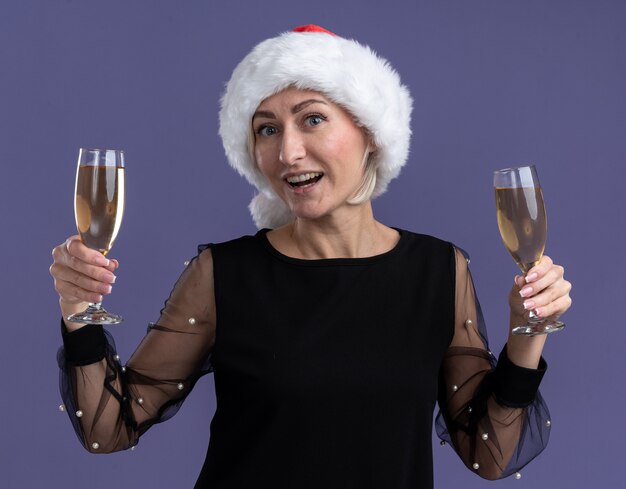 Gioiosa donna bionda di mezza età che indossa il cappello di Natale che guarda l'obbiettivo che tiene due bicchieri di champagne isolato su sfondo viola
