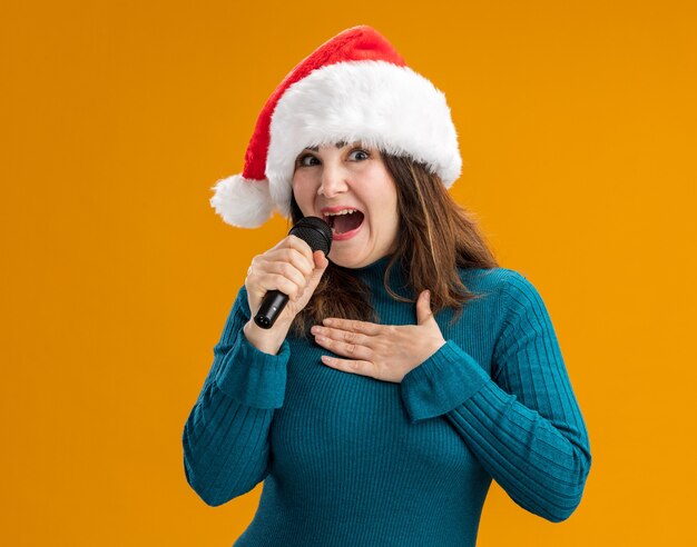 gioiosa donna caucasica adulta con cappello santa tiene il microfono e mette la mano sul petto isolato su sfondo arancione con spazio di copia