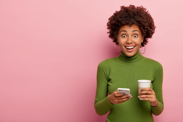 Gioiosa donna dai capelli ricci con pelle sana, indossa un turtlenek verde casual, detiene un moderno caffè cellulare e da asporto, gode della comunicazione online