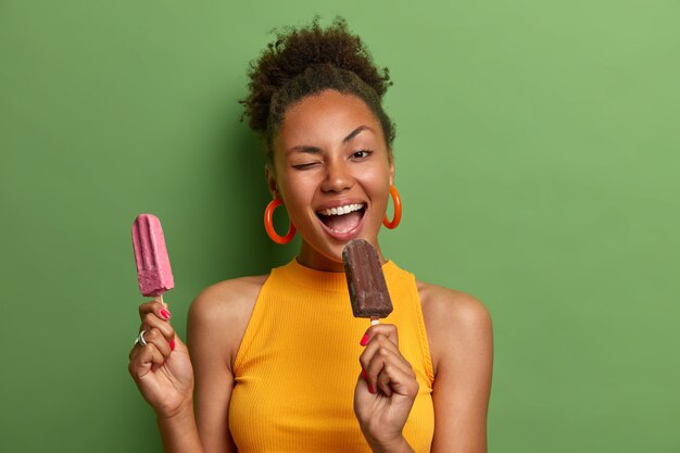 Gioiosa donna dai capelli ricci morde delizioso gelato strizza l'occhio ha divertimento felice espressione