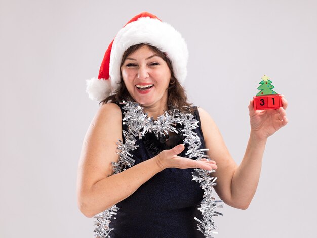 Gioiosa donna di mezza età che indossa cappello da Babbo Natale e ghirlanda di orpelli intorno al collo tenendo e puntando al giocattolo dell'albero di Natale con data guardando la fotocamera isolata su sfondo bianco