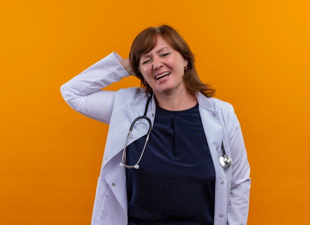 Gioiosa donna di mezza età medico indossa abito medico e stetoscopio mettendo la mano dietro la testa sulla parete arancione isolata