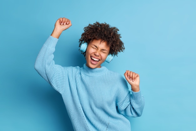 Gioiosa donna riccia dalla pelle scura balla spensierata alza le braccia si sente ottimista indossa le cuffie stereo gode della musica preferita indossa un maglione lavorato a maglia