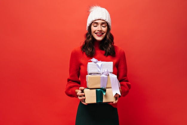 Gioiosa donna riccia in maglione rosso brillante, cappello a maglia posa gli occhi chiusi e tiene scatole regalo su un muro rosso isolato isolated