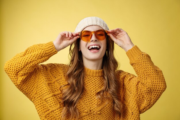 Gioiosa elegante ragazza caucasica che si gode la luce del sole stazione sciistica invernale indossando occhiali da sole cappello maglione caldo divertendosi sorridente toccando occhiali, esprimendo umore giocoso felice sfondo giallo