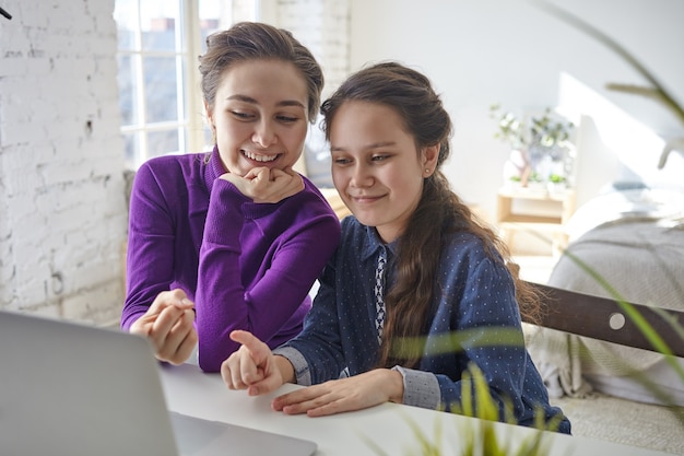 Gioiosa felice giovane madre e figlia shopping online utilizzando il pc portatile, seduto alla scrivania all'interno della camera da letto leggera, puntando le dita sullo schermo e sorridente