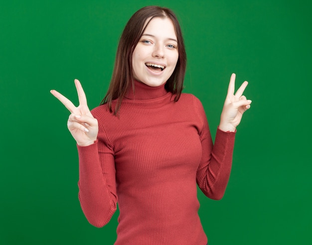 Gioiosa giovane bella donna che guarda davanti facendo segno di pace isolato sul muro verde