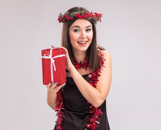 Gioiosa giovane bella ragazza caucasica che indossa ghirlanda di testa di Natale e ghirlanda di orpelli intorno al collo che tiene il pacchetto regalo guardando la fotocamera isolata su sfondo bianco con spazio di copia