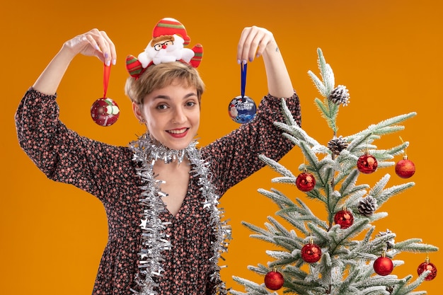 gioiosa giovane bella ragazza che indossa la fascia di babbo natale e la ghirlanda di orpelli intorno al collo in piedi vicino all'albero di natale decorato che tiene la pallina di natale vicino alla testa isolata sul muro arancione