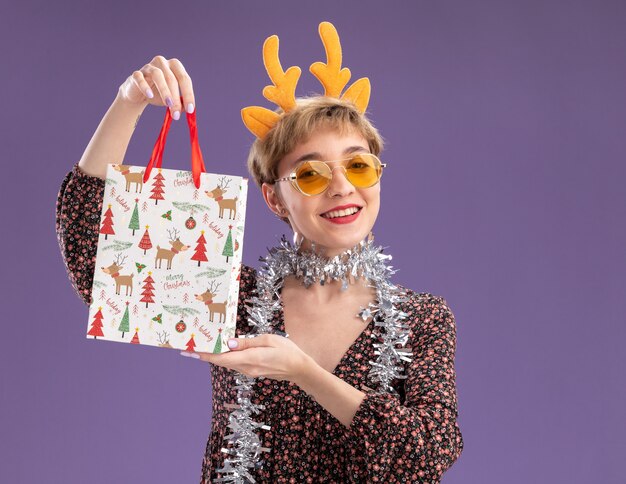 Gioiosa giovane bella ragazza che indossa la fascia di corna di renna e la ghirlanda di orpelli intorno al collo con gli occhiali che tengono il sacchetto del regalo di natale isolato sulla parete viola con lo spazio della copia