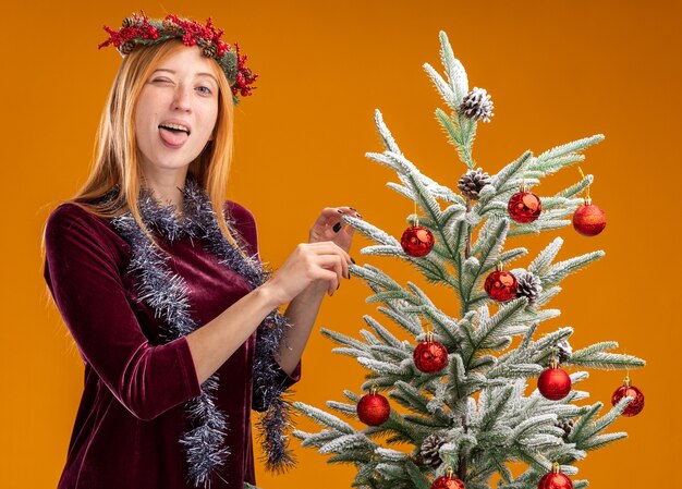 Gioiosa giovane bella ragazza in piedi vicino all'albero di Natale che indossa un abito rosso e la corona con la ghirlanda sul collo che mostra la linguetta isolata su sfondo arancione