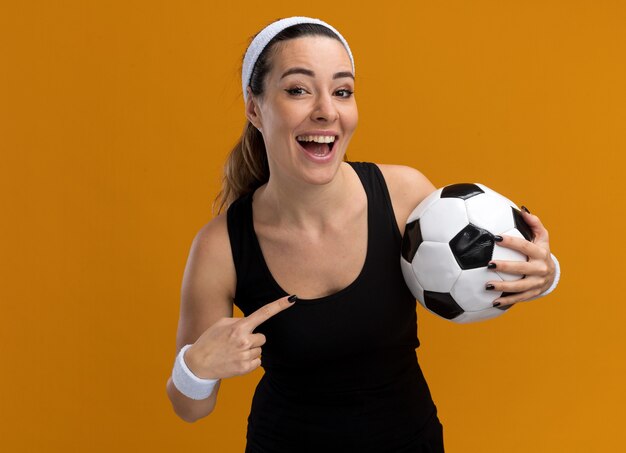 Gioiosa giovane donna abbastanza sportiva che indossa fascia e braccialetti che tengono il pallone da calcio che lo punta guardando davanti isolato sul muro arancione con spazio di copia