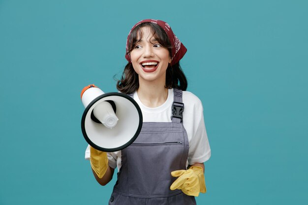 Gioiosa giovane donna addetta alle pulizie che indossa bandana uniforme e guanti di gomma che tengono l'altoparlante guardando il lato isolato su sfondo blu