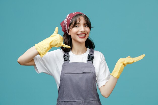 Gioiosa giovane donna addetta alle pulizie che indossa bandana uniforme e guanti di gomma guardando la fotocamera che mostra la mano vuota facendo un gesto di chiamata isolato su sfondo blu