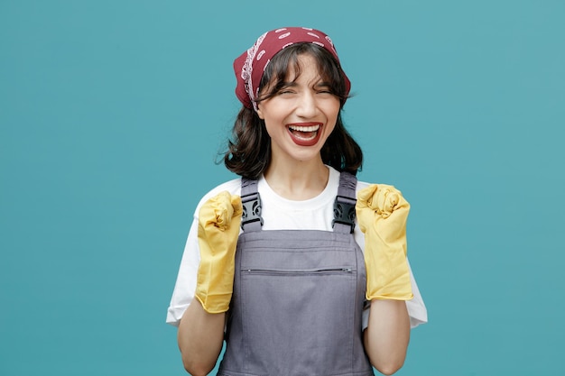 Gioiosa giovane donna addetta alle pulizie che indossa bandana uniforme e guanti di gomma guardando la fotocamera che mostra sì gesto isolato su sfondo blu