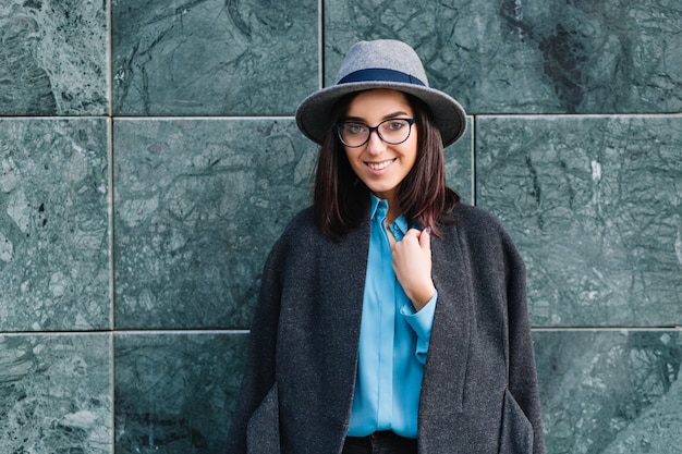 Gioiosa giovane donna alla moda in cappello grigio, cappotto, occhiali neri sorridente e in posa sul muro grigio. Abiti di lusso, modello alla moda, umore allegro.