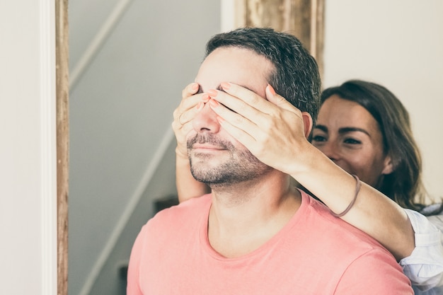 Gioiosa giovane donna che copre gli occhi del suo ragazzo con le mani e lo conduce nel loro nuovo appartamento.